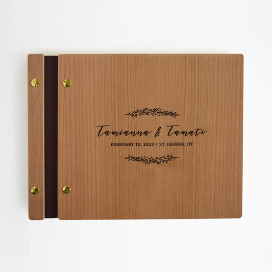 An 8.5x11" guest book lies on a table. Made of cherry wood, black vegan leather binding, and copper hardware. The front cover includes an engraved design with personalized names. 
