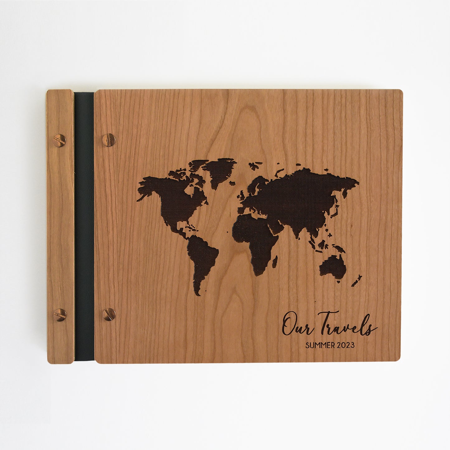 An 8.5x11" guest book lies on a table. Made of cherry wood, black vegan leather binding, and copper hardware. The front cover has en engraved world map and reads Our Travels, Summer 2023.