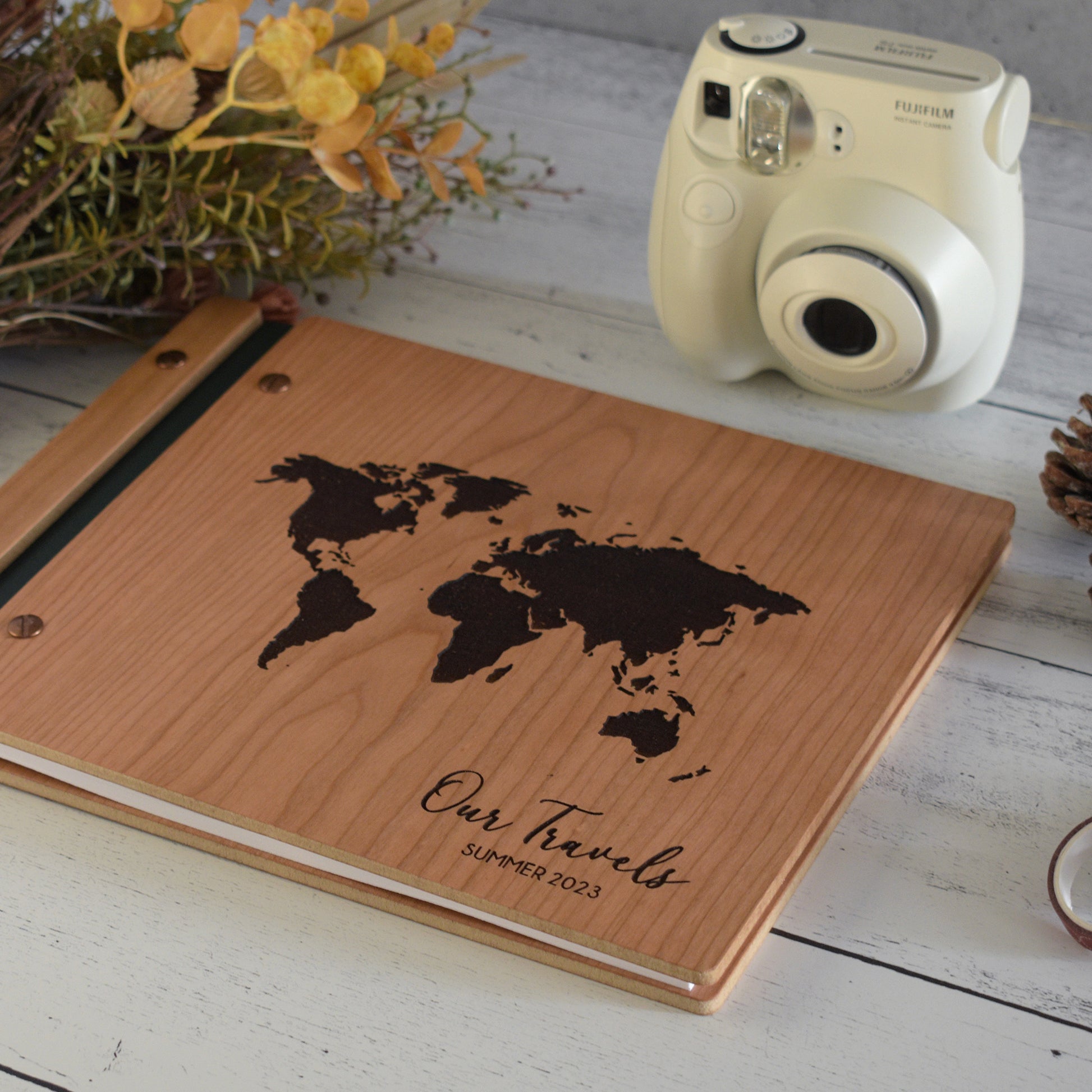 An 8.5x11"guest book lies on a table. Made of cherry wood, black vegan leather binding, and copper hardware. The front cover has en engraved world map and reads Our Travels, Summer 2023.