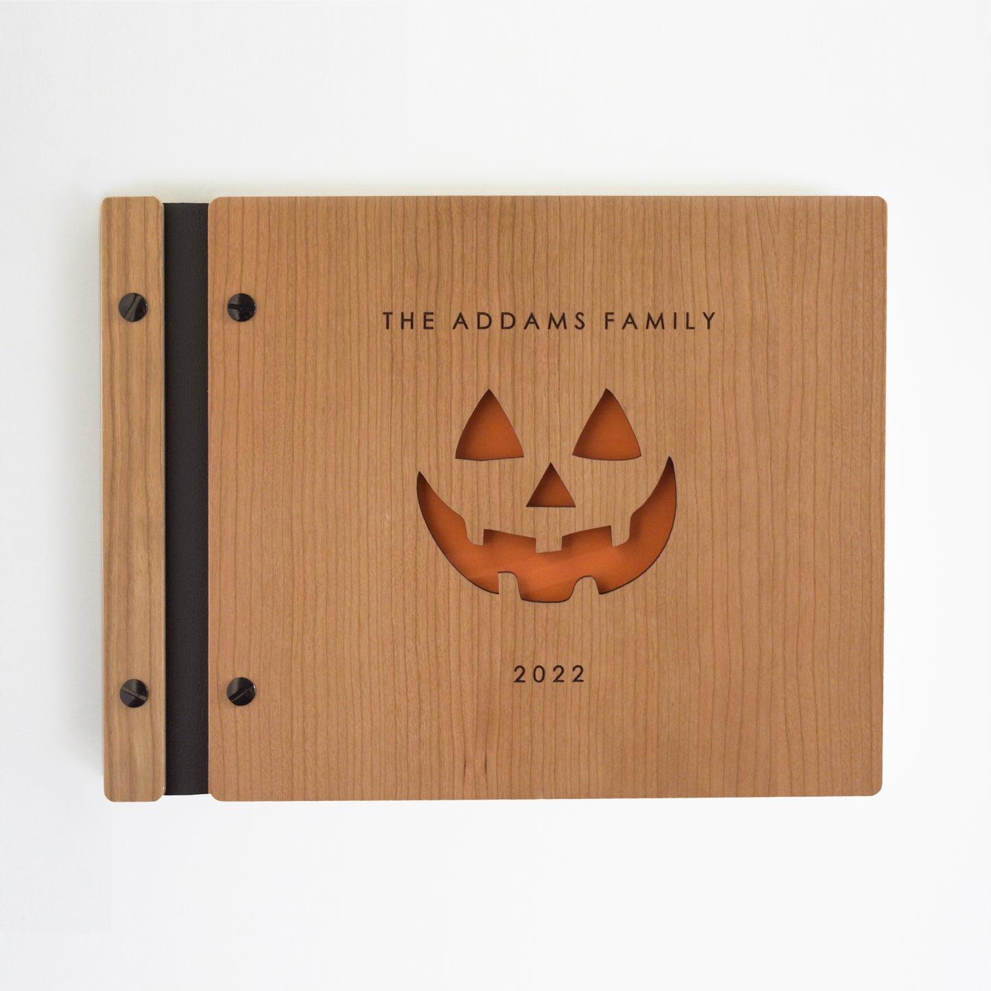 An 8.5x11" guest book lies on a table. Made of cherry wood, black vegan leather binding, and black hardware. The front cover reads The Addams Family, 2022 with an engraved pumpkin in the center.
