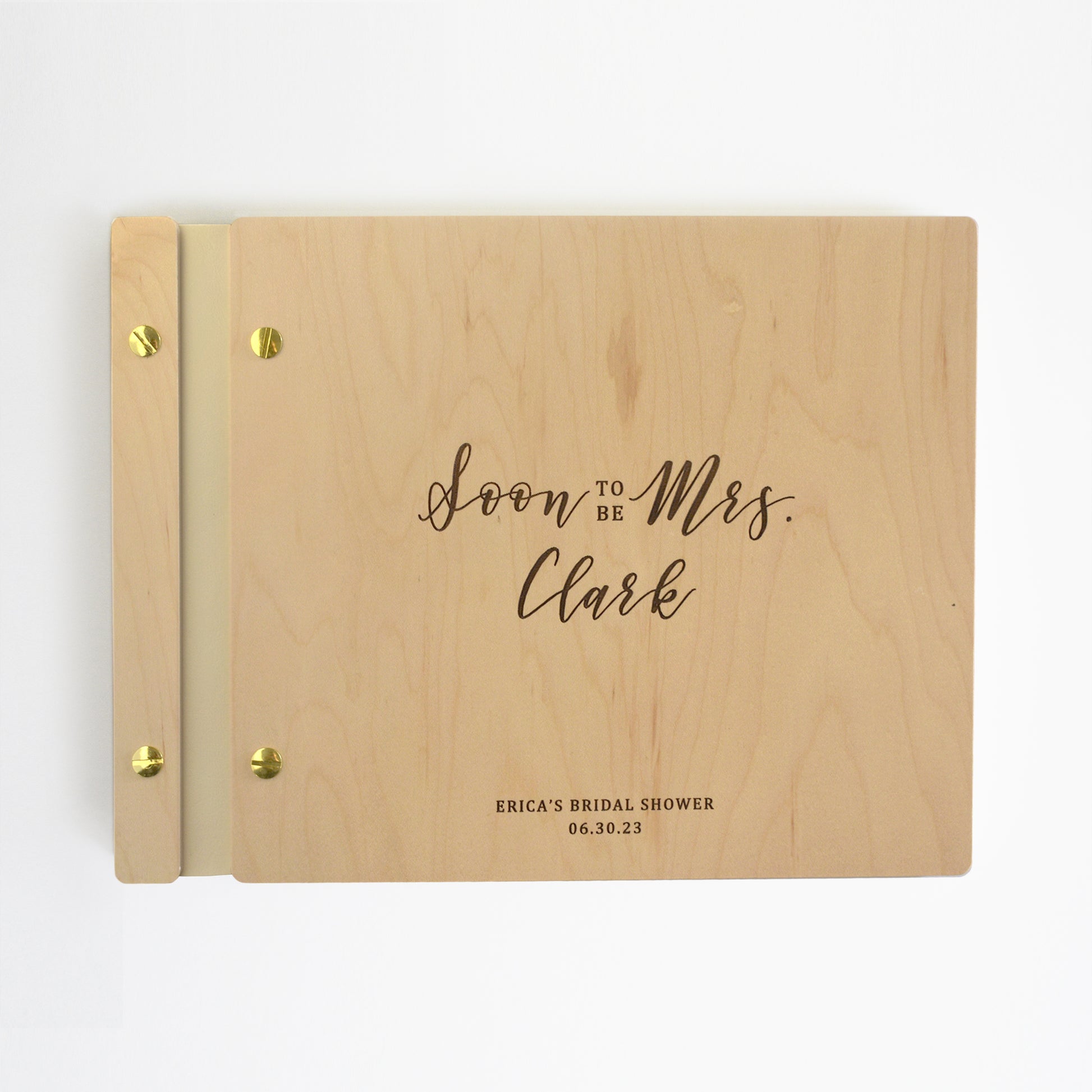 An 8.5x11" wedding guest book lies on a table. Made of cherry or maple wood, vegan leather binding, and aluminum hardware. The front cover includes an engraved design with personalization.