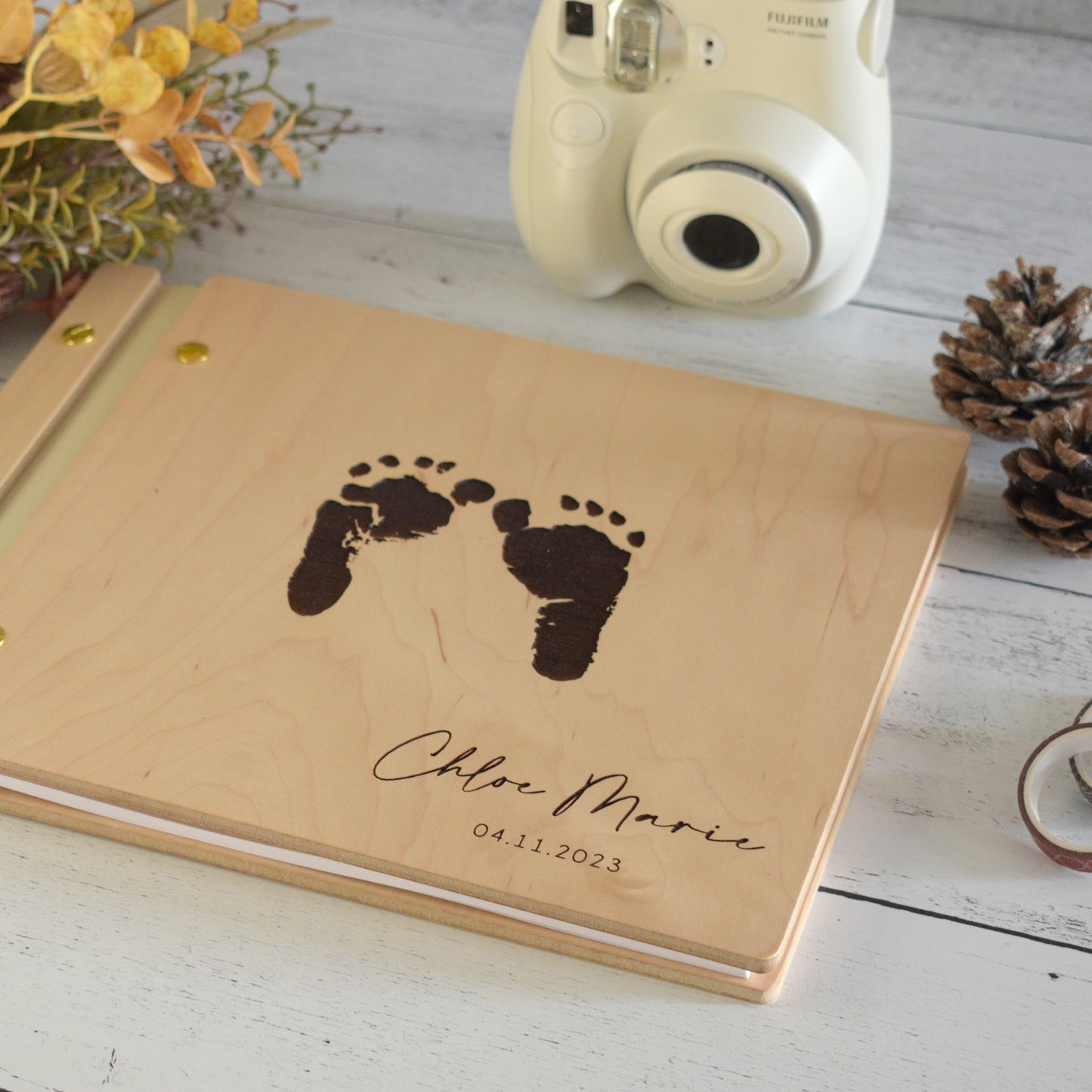 An 8.5x11" guest book lies on a table. Made of maple wood, ivory vegan leather binding, and gold hardware. The front cover has engraved baby foot prints with the words Chloe Marie 04.11.2023.