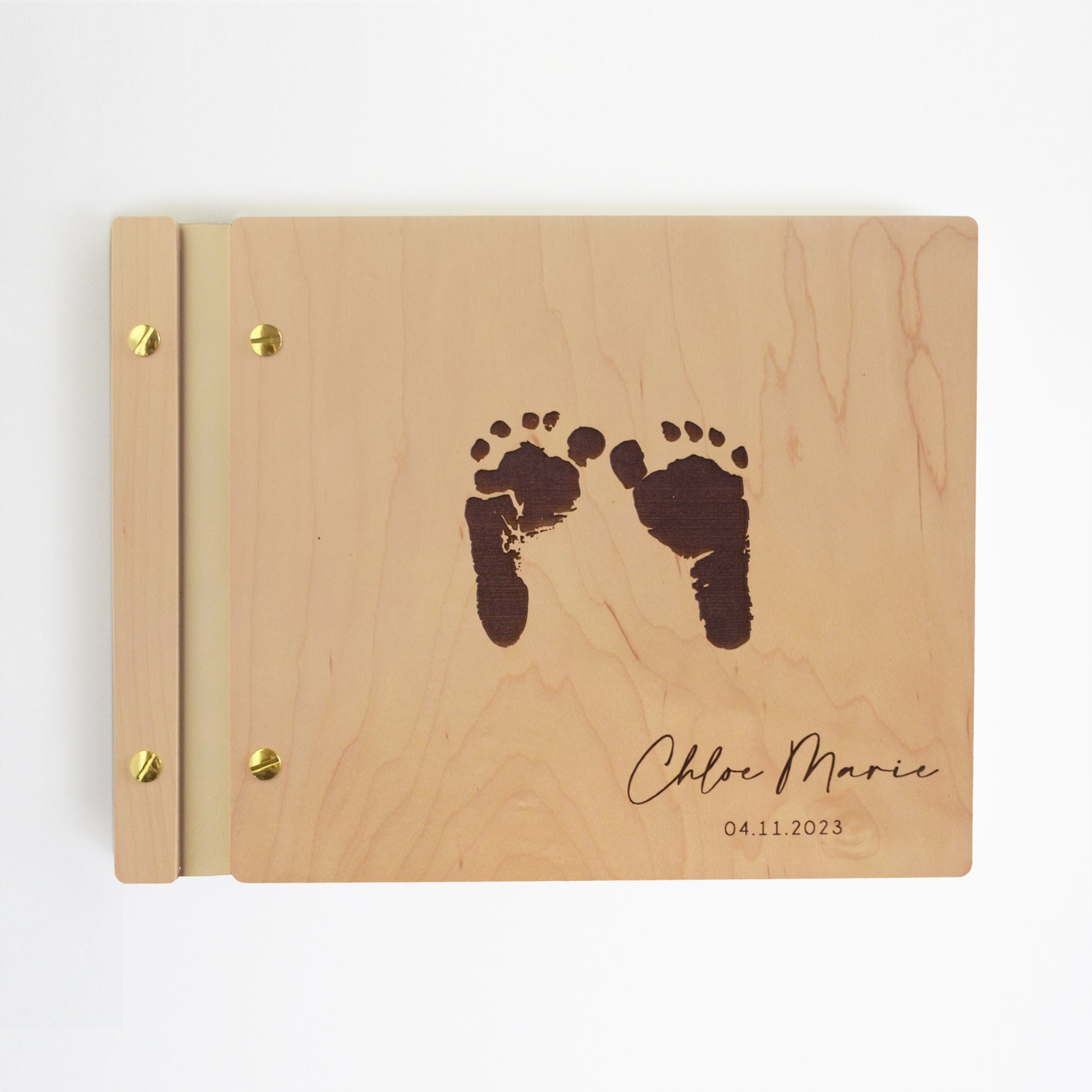 An 8.5x11" guest book lies on a table. Made of maple wood, ivory vegan leather binding, and gold hardware. The front cover has engraved baby foot prints with the words Chloe Marie 04.11.2023.