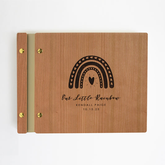 An 8.5x11" guest book lies on a table. Made of cherry wood, gold vegan leather binding, and gold hardware. The front cover reads Our Little Rainbow, Kendall Paige, 10.15.2023 with an engraved rainbow.