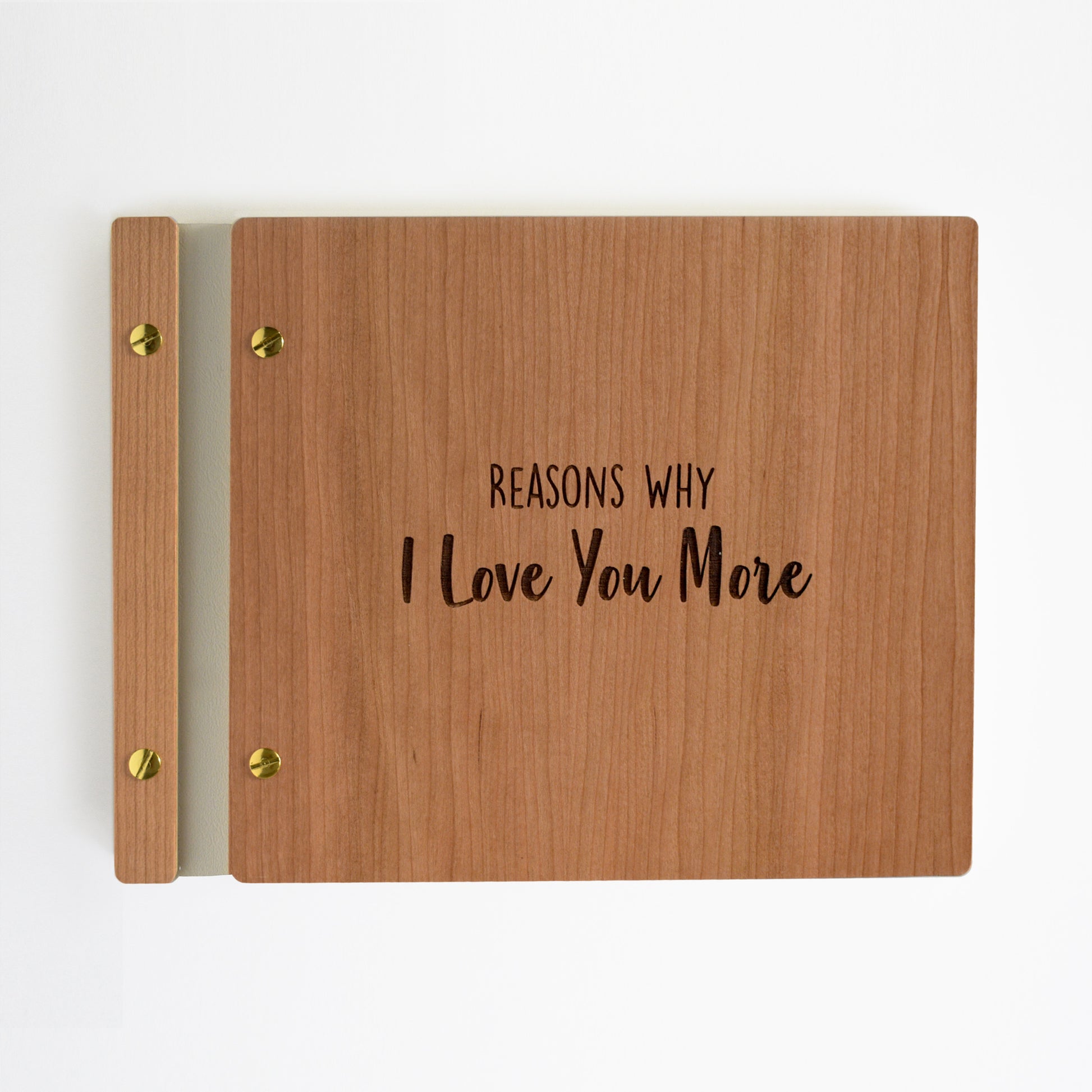 An 8.5x11" guest book lies on a table. Made of cherry wood, ivory vegan leather binding, and gold hardware. The front cover reads “Reasons Why I Love You More.”