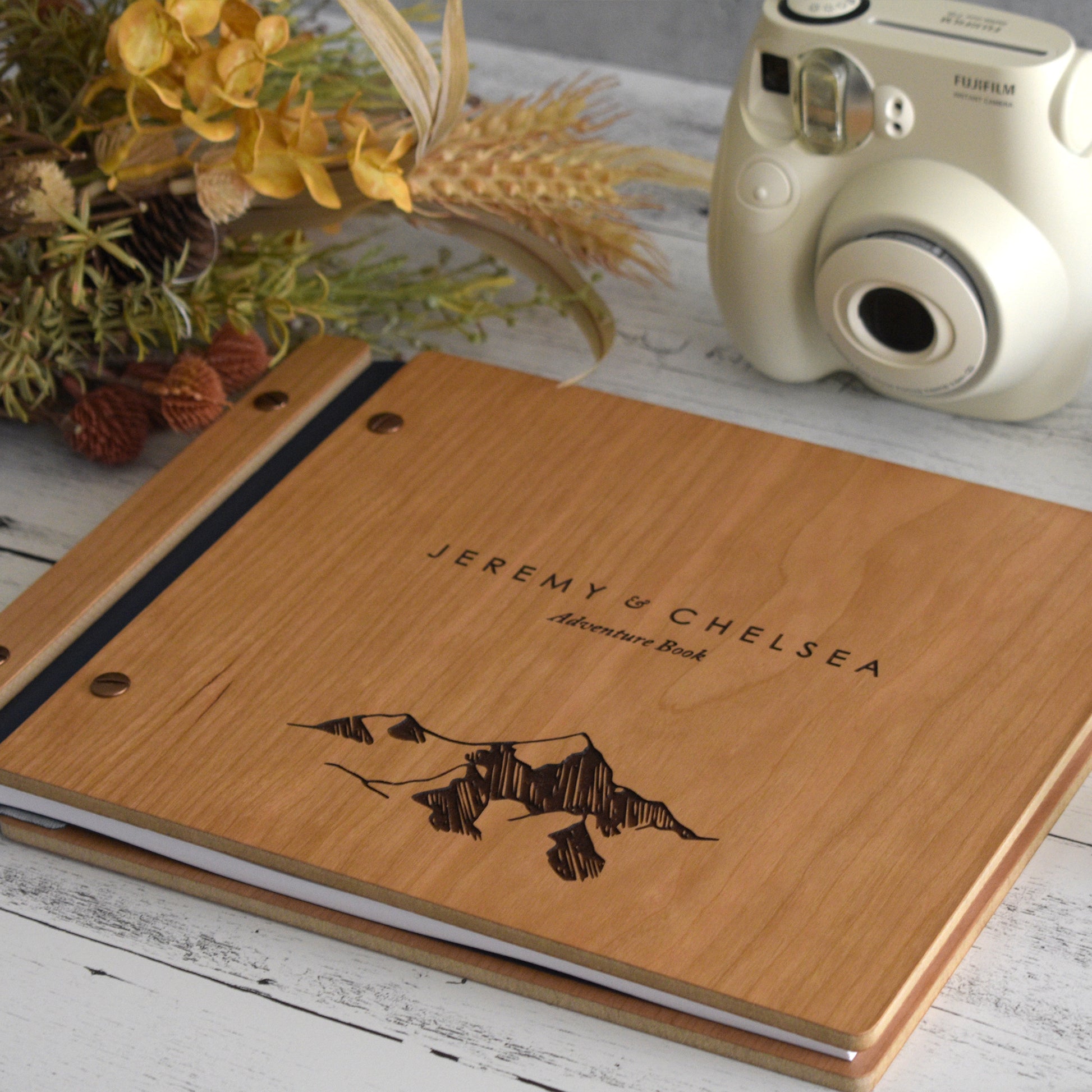 An 8.5x11" guest book lies on a table. Made of cherry wood, black vegan leather binding, and copper hardware. The front cover has an engraved mountain image and reads Jeremy & Chelsea, Adventure Book.