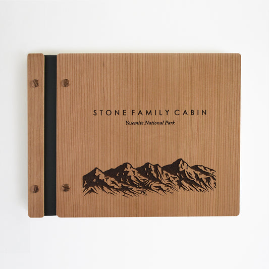 An 8.5x11" Airbnb guest book lies on a table. Made of cherry wood, black vegan leather binding, and copper hardware. The front cover reads Stone Family Cabin, Yosemite National Park.