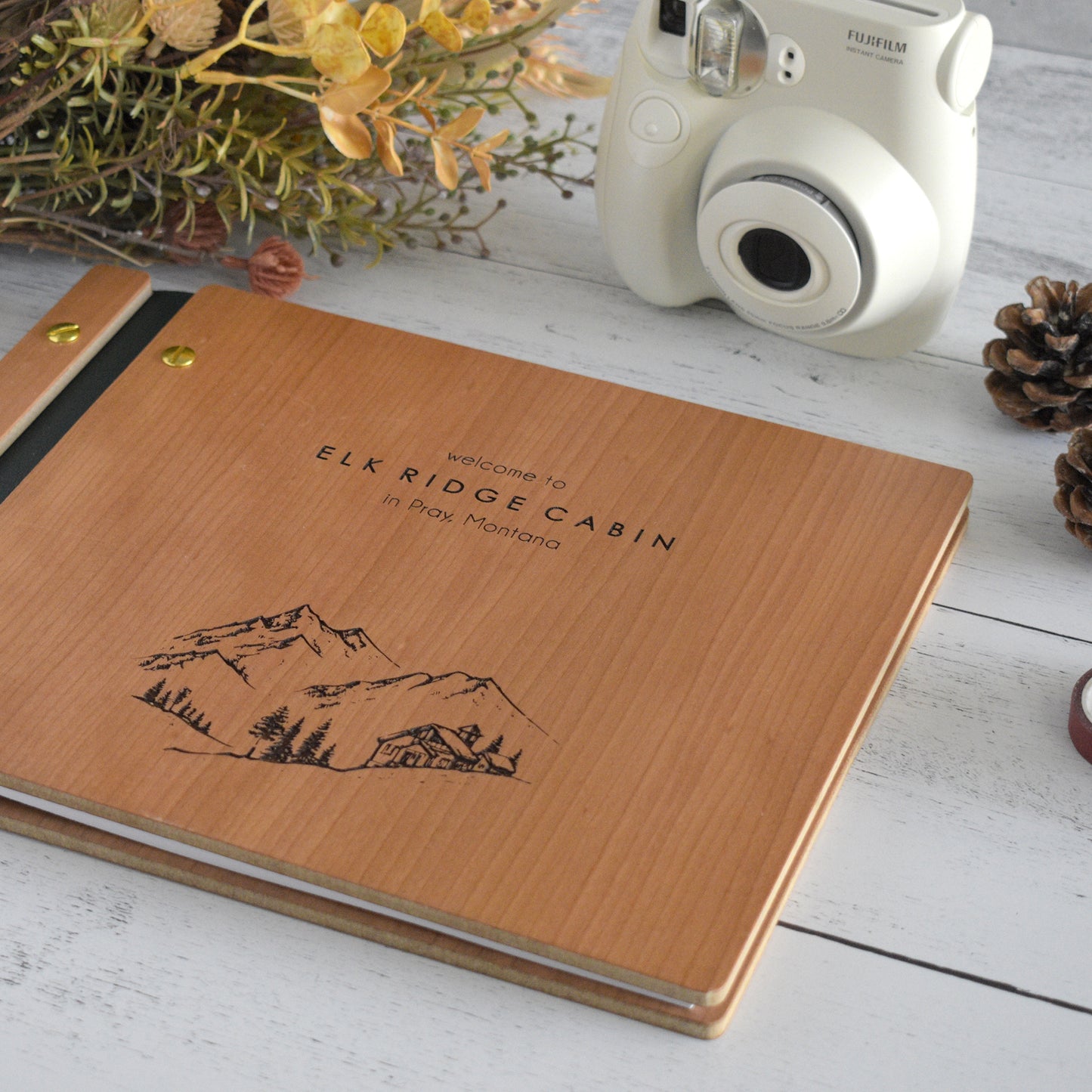 An 8.5x11" Airbnb guest book lies on a table. Made of cherry wood, black vegan leather binding, and gold hardware. The front cover reads Welcome to Elk Ridge Cabin in Pray, Montana.