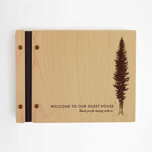 An 8.5x11" Airbnb guest book lies on a table. Made of maple wood, black vegan leather binding, and copper hardware. The front cover reads Welcome to our guest house, Thank you for staying with us.