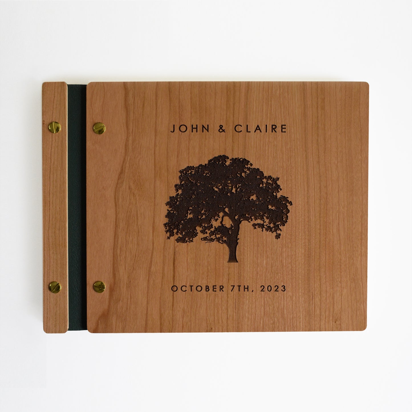An 8.5x11" guest book lies on a table. Made of cherry wood, black vegan leather binding, and copper hardware. The front cover includes an engraved design with personalized names.
