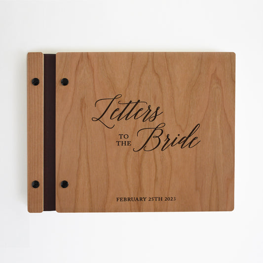 An 8.5x11" guest book lies on a table. Made of cherry wood, black vegan leather binding, and black hardware. The front cover includes an engraved design with personalization.