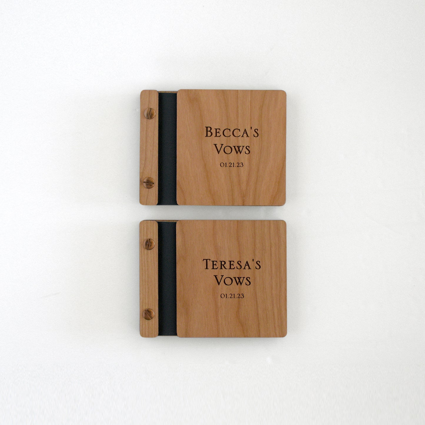 Two vow books sit on a table next to one another. Made of maple wood, aqua binding, and silver hardware, they read Becca's Vows and Teresa's Vows.