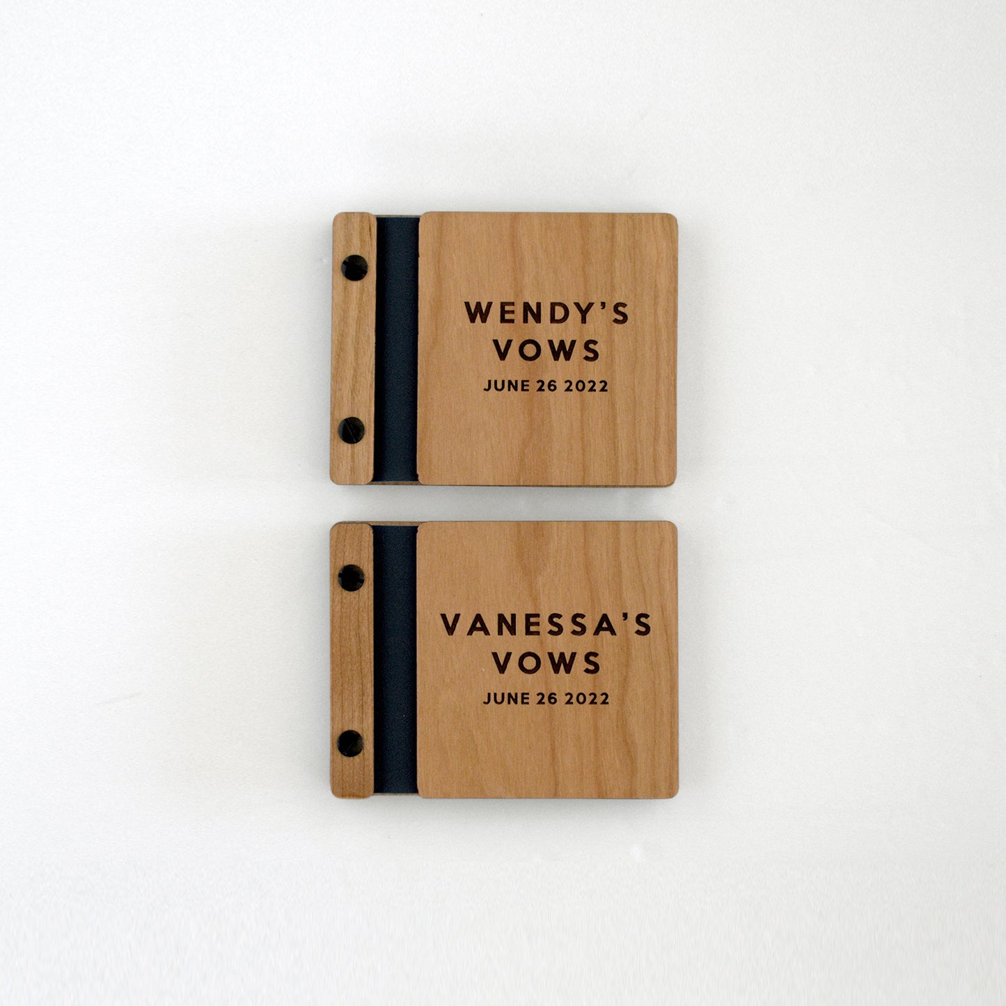 Two vow books sit on a table next to one another. Made of cherry wood, black binding, and black hardware, they read Wendy's Vows, Vanessa's Vows.