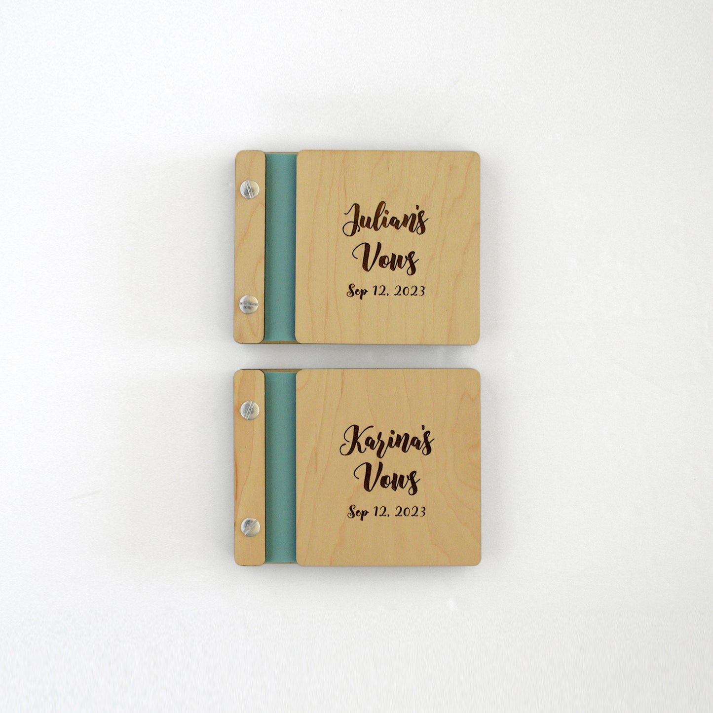 Two vow books sit on a table next to one another. Made of maple wood, aqua binding, and silver hardware, they read Julian’s Vows and Karina’s Vows.