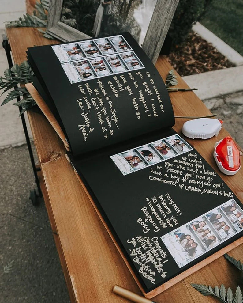 An 8.5x11 inch guest book lies open on a wooden desk. Black cardstock pages with handwritten messages and photo booth strips are posted on the pages for the bride and groom to cherish as a keepsake after their wedding day.