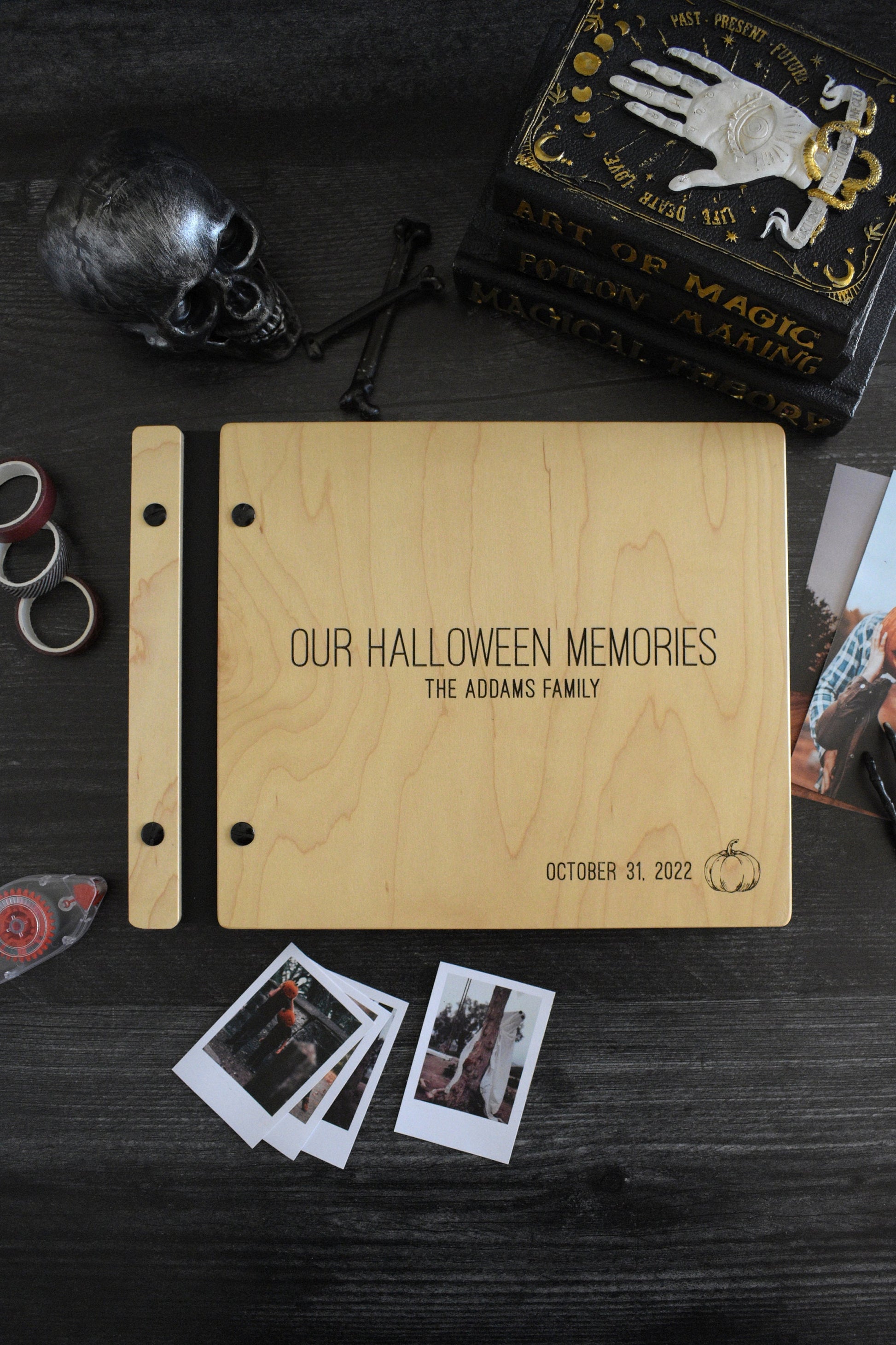 An 8.5x11" guest book lies on a table. Made of maple wood, black vegan leather binding, and black hardware. The front cover reads Our Halloween Memories, The Addams Family, October 31, 2022 with an engraving of a pumpkin in the bottom right corner of the book.