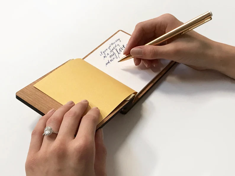 A vow book lies open as a female writes on the white cardstock pages with a gold pen.
